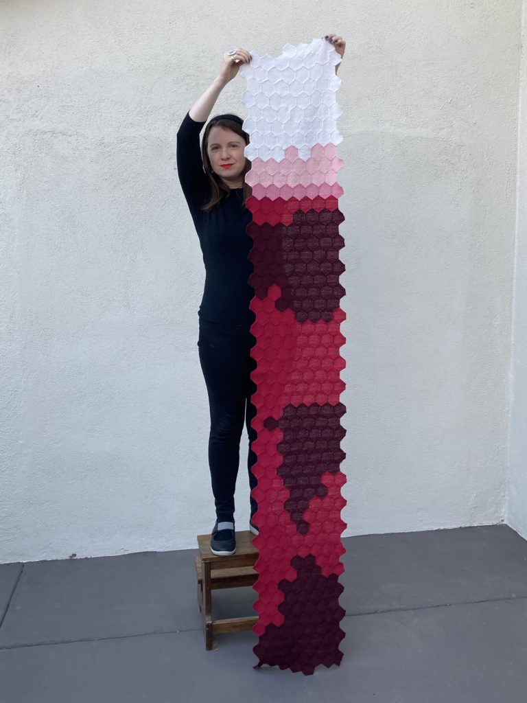 Woman holding a long scarf vertically. Scarf is made up of tiny woven hexagons sewn together. Hexagons in the top are white, then progress through pinks to red. There are three large blotches of dark red within the large red section.