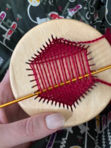 Small hexagon pin loom, half woven on top and bottom with red yarn.