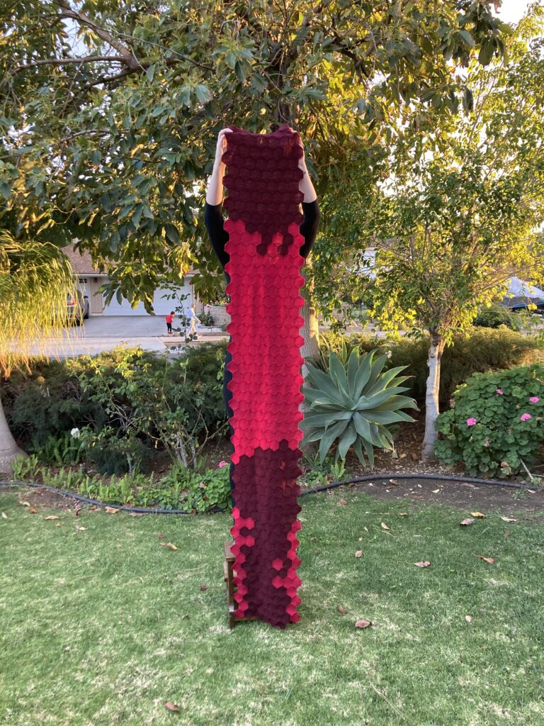 Person hidden behind a narrow vertical piece of fabric made up of small red and dark red hexagons. The top quarter of the fabric is dark red, with red hexagons through the middle, and mostly dark red in bottom third. In the background is a green yard with plants and bushes.