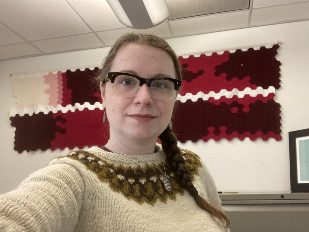 White woman with glasses and braided brown hair wearing a colorwork-yoked sweater stands in front of the picture as a selfie. In the background, two long narrow strips of fabric hang on the white wall behind her. The fabric is made up of small hexagons in colors ranging from white and pink to red and dark red.