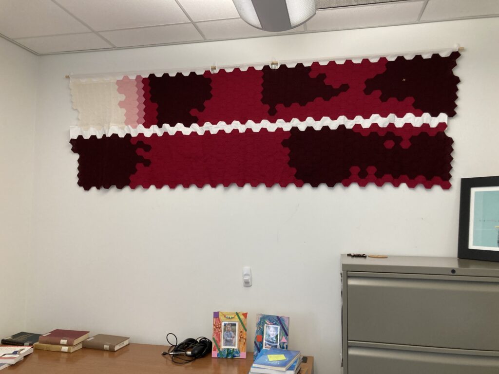 White wall of an office with two long fabric strips hanging toward the top of the wall, one strip on top of the other. The top strip is thin and long, starting white on the left and transitioning to red, with three large dark red patches. The lower strip hangs directly below the first and is dark red on the ends and red in the middle. Other office furniture, art, and books are visible.